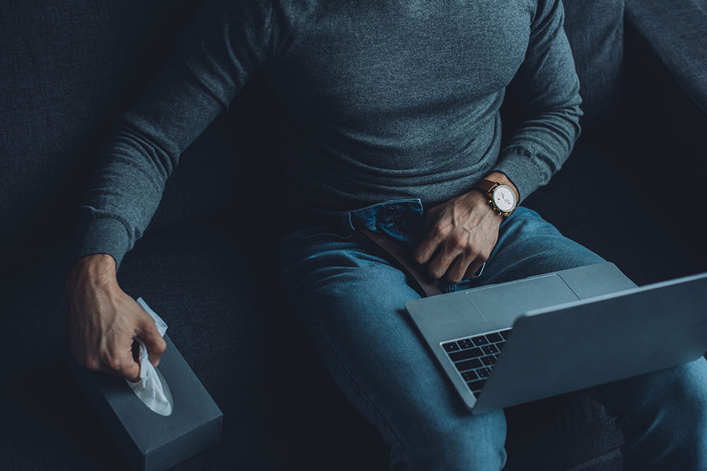 Mann mit geöffneter Hose vor Notebook, eine Hand im Intimbereich, andere Hand in der Taschentuch-Box