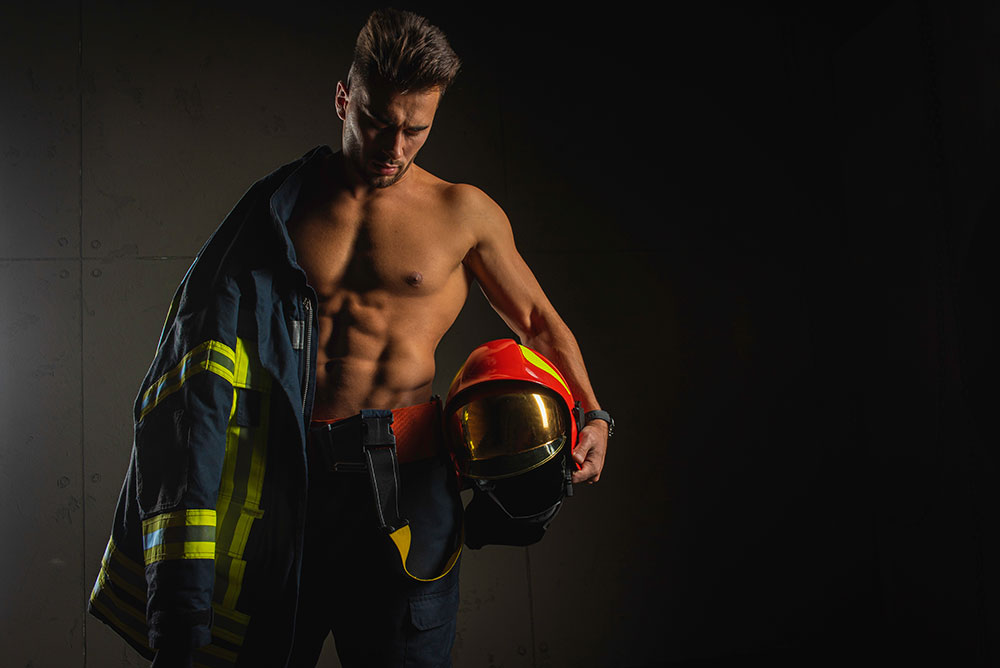 Mann mit nacktem Oberkörper und Feuerwehrjacke über Schulter und Helm unter dem Arm